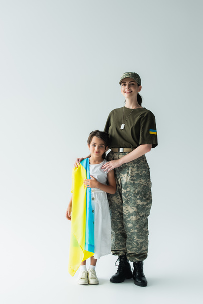 Full length of smiling woman in camouflage uniform hugging daughter with ukrainian flag on grey background - Фото, изображение