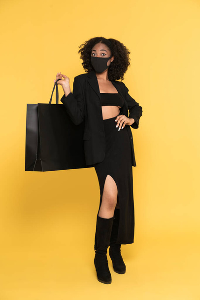 Black woman in face mask posing with shopping bag isolated over yellow background - Fotó, kép