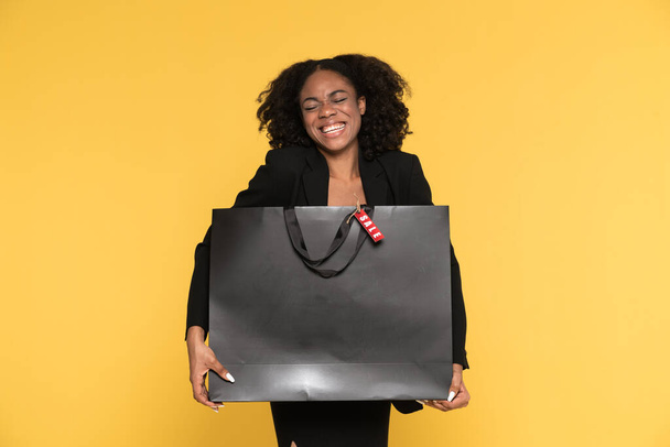 Чорна жінка сміється під час позування з сумкою для покупок ізольовано на жовтому тлі
 - Фото, зображення