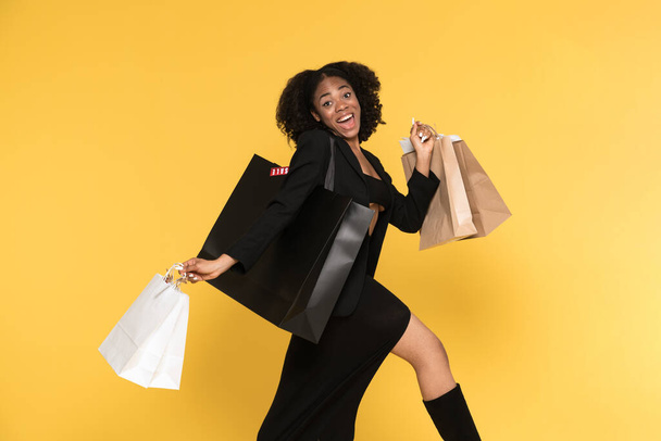 Young black woman laughing while walking with shopping bags isolated over yellow background - Foto, Imagem