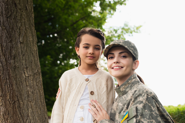 Smiling soldier with ukrainian flag on chevron hugging daughter outdoors  - Foto, Bild