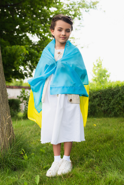 full length of patriotic child covered with ukrainian flag in park - 写真・画像