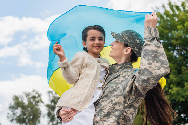 cheerful military woman with daughter holding ukrainian flag outdoors - Foto, immagini