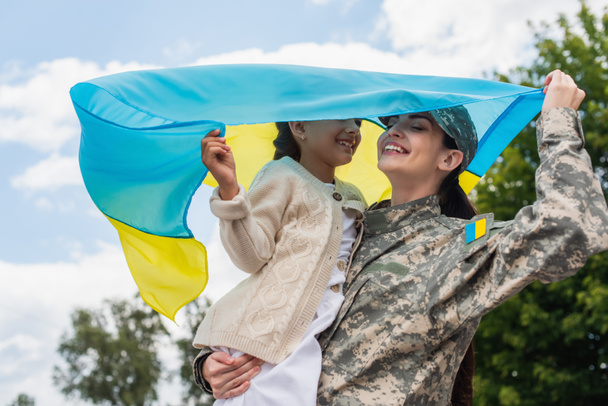 smiling woman in camouflage holding ukrainian flag with daughter outdoors - Foto, Imagem