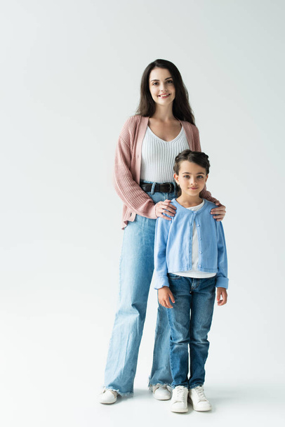 full length of positive woman with daughter in jeans standing on grey background - Valokuva, kuva