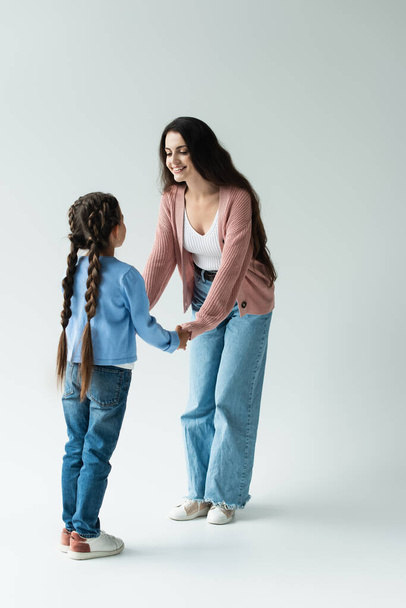 full length of cheerful woman holding hands of daughter with pigtails on grey background - Foto, Bild