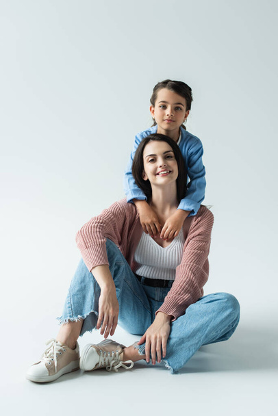 child looking at camera and embracing mother sitting in jeans on grey background - Photo, Image
