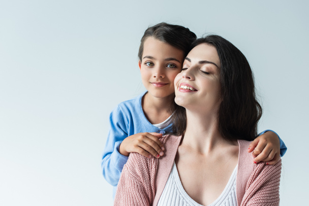 brunette woman with closed eyes smiling near daughter looking at camera isolated on grey - Foto, Imagem