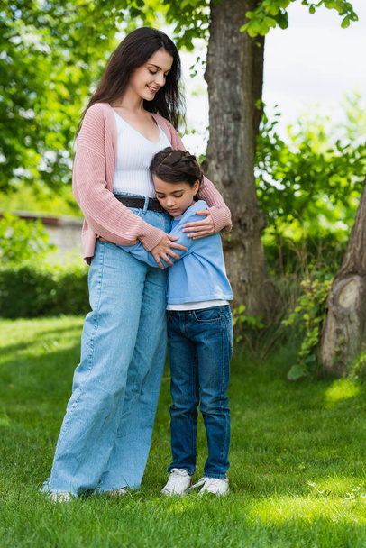 pleased woman in jeans embracing daughter in park - Фото, изображение
