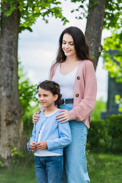 smiling woman embracing shoulders of pensive daughter in park - Zdjęcie, obraz