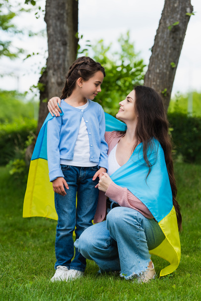 mam en dochter bedekt met Oekraïense vlag glimlachen naar elkaar in het park - Foto, afbeelding
