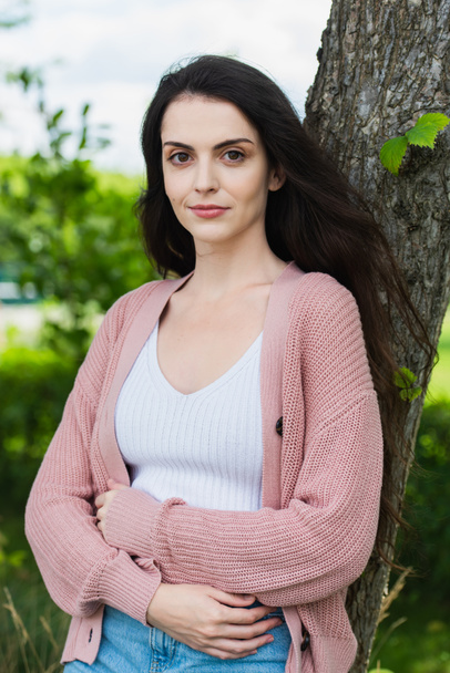 positive brunette woman standing with crossed arms in park and looking at camera - Fotografie, Obrázek