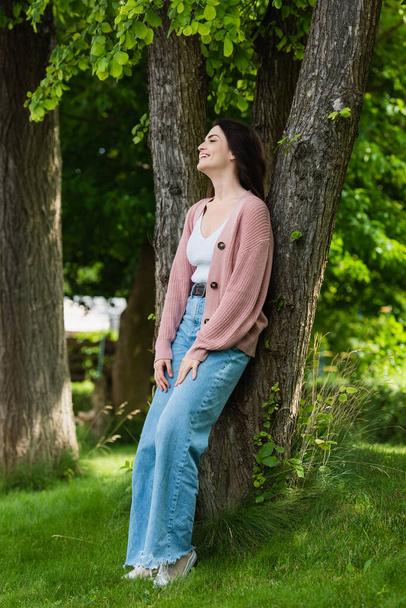 brunette woman in jeans smiling near trees in park - Photo, image