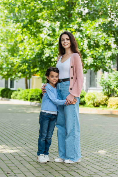 full length of mom and daughter in jeans embracing and smiling at camera on city street - Foto, immagini