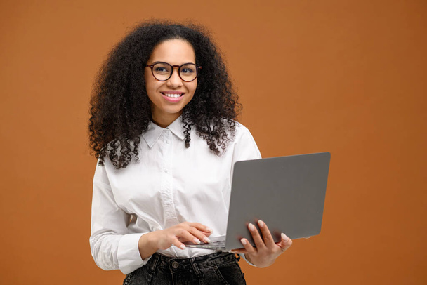 Smart and confident African-American woman in smart casual wear using laptop isolated on brown background, female office employee or student typing, web browsing - Zdjęcie, obraz
