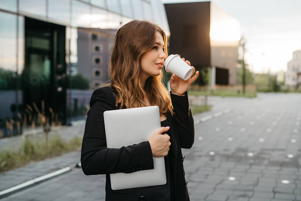Lifestyle portrait of a young and cheerful woman standing with phone and coffee cup outdoors. Urban business travel and transportation concept - Fotó, kép