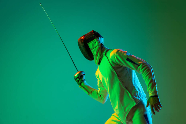 Studio shot of young man, fencer with smallsword practicing fencing isolated on green background in neon light. Athlete practicing in motion, action. Copyspace for ad. - Photo, Image