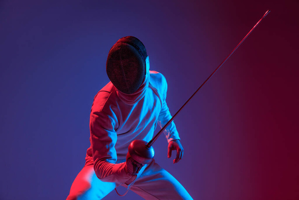 Fight in swordplay. Male fencer with smallsword practicing fencing isolated on purple background in neon light. Athlete practicing in motion, action. Copyspace for ad. - Фото, изображение