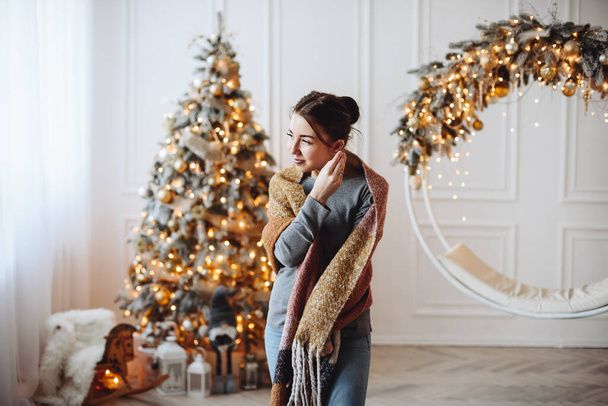 Young Woman is Standing in Warm Clothes and Scarf in Room Against Backdrop of Christmas Tree Decorated with Golden Lights. Atmosphere of Magic - Foto, Bild