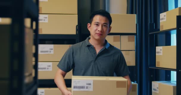 Успішний азіатський бізнесмен стоїть навпроти полиці і тримає картонні коробки з паперу, посміхаючись, і дивиться на камеру, горду тим, що вночі презентує свій бізнес на складі. Початкова концепція малого бізнесу. - Кадри, відео