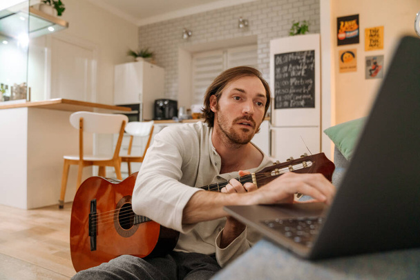 Ginger man playing guitar and using laptop while sitting on floor at home - Foto, Imagen
