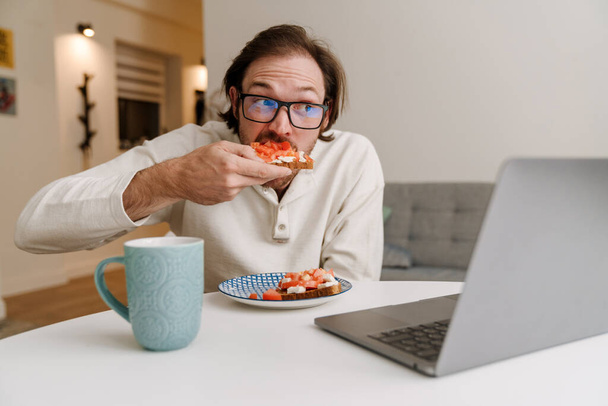 Ginger bristle man in eyeglasses working with laptop while having dinner at home - Fotó, kép