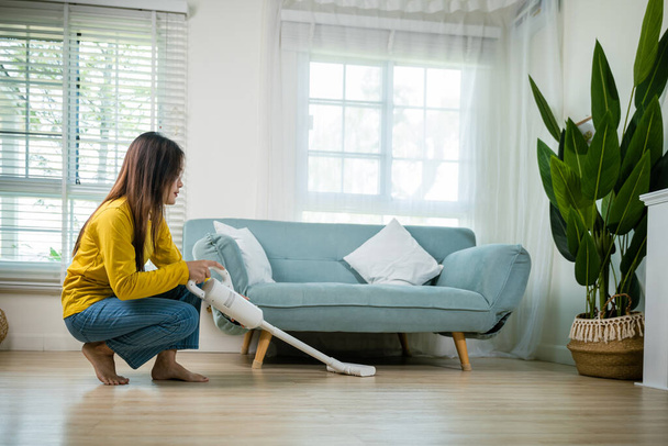 Housewife female dust cleaning floor under sofa or couch furniture with vacuum cleaner, Happy Asian young woman with accumulator vacuum cleaner at home in living room, household and housework concept - Foto, afbeelding