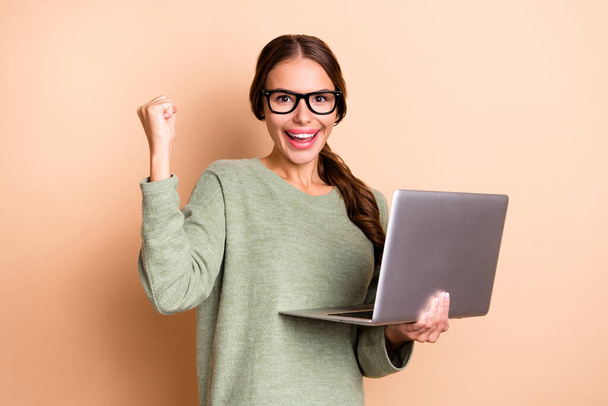Photo of excited lucky lady wear green sweater spectacles rising fist working modern gadget isolated beige color background. - Фото, изображение