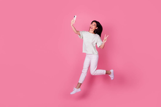 Photo of cute young japanese lady hold gadget take selfie jump show v-sign dressed stylish white clothes isolated on pink color background. - Valokuva, kuva