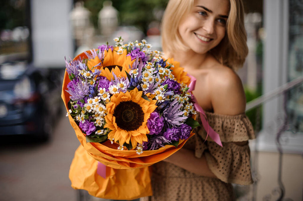 smiling young woman holds bouquet of bright yellow sunflowers and daisies and purple flowers packed in wrapping paper in her hands - Fotoğraf, Görsel