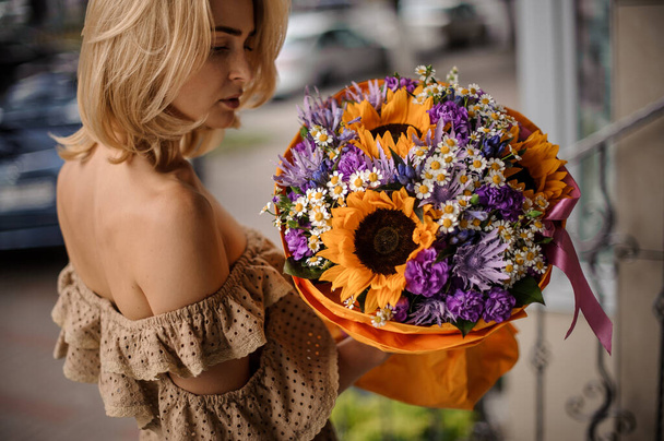 beautiful woman holds bouquet of bright yellow sunflowers and daisies and purple flowers packed in wrapping paper in her hand - Fotoğraf, Görsel