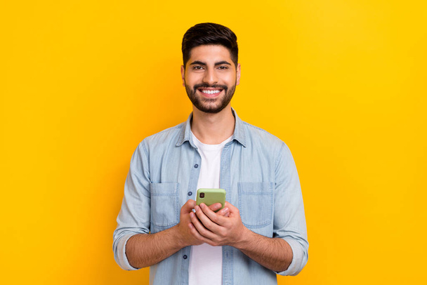 Portrait of young happy man wear denim shirt businessman got bitcoin profit holding phone isolated on yellow color background. - Фото, изображение