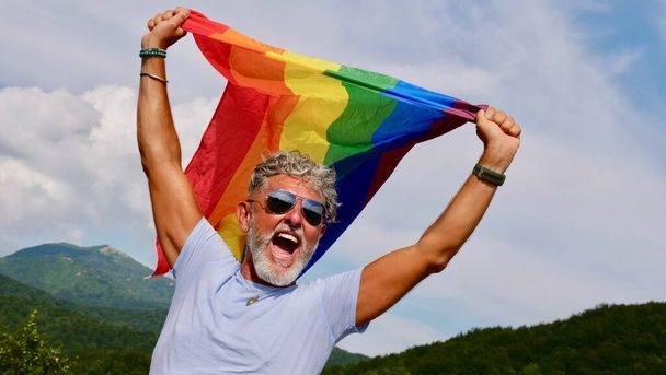 Portret van een grijze bejaarde blanke man met een baard en een zonnebril met een regenboog LGBTQIA vlag tegen een achtergrond in de lucht, schreeuwt uit protest, viert Pride Month Coming Out Day - Foto, afbeelding