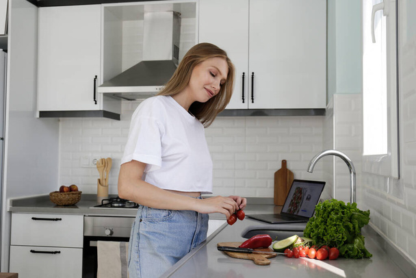 Young beautiful woman alone at the kitchen with white cupboard and tiling, cooking a salad. Portrait of a female chopping the ingredients for the vegan dish. Close up, copy space, background. - Foto, Bild