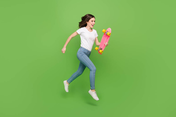 Photo of impressed funky young lady wear white t-shirt jumping running riding long board isolated green color background. - Фото, зображення
