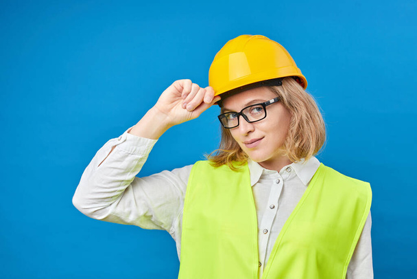 Young woman engineer wearing a yellow helmet, standing in studio on blue background. The concept of construction, repair, decoration - Fotó, kép