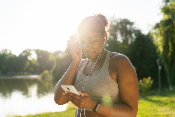 Plus grote Afro-Amerikaanse vrouw met behulp van mobiele telefoon en oortelefoons voor het sporten in het park in een zomerdag  - Foto, afbeelding