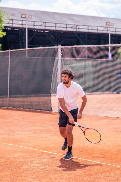 Great looking man professional tennis player hitting hard the tennis ball with the racket while playing the match outdoor on the clay court. - Foto, Imagen