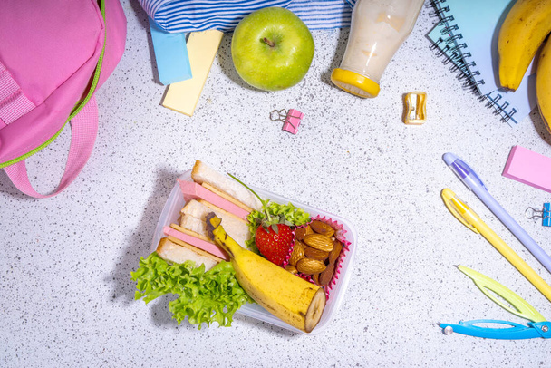 Almuerzo escolar saludable: sándwich, verduras, frutas, nueces y yogur con útiles escolares, accesorios y mochila en el espacio de copia plana de fondo negro. Regreso al concepto escolar - Foto, Imagen