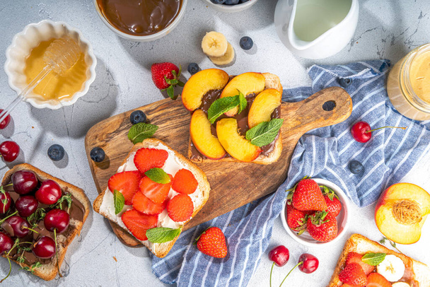  American European summer breakfast sandwiches, toasts with peanut butter, chocolate and cream cheese with berry and fruits, top view flatlay on kitchen table - Foto, Imagem