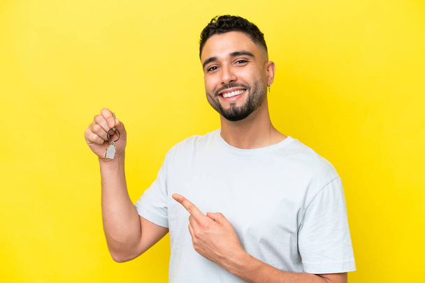 Young Arab man holding home keys isolated on yellow background and pointing it - Foto, Imagem