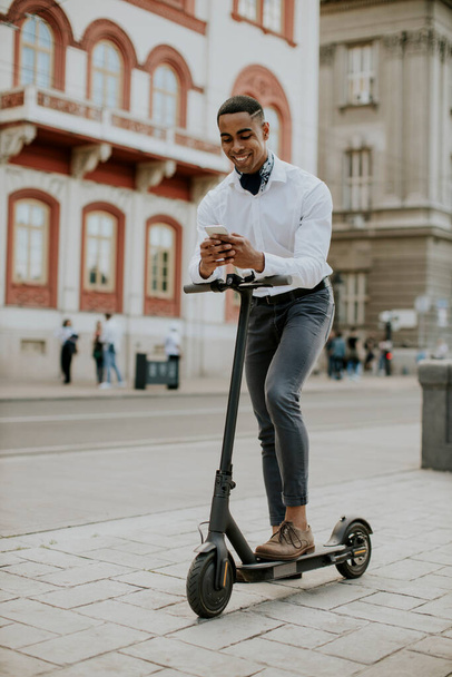 Beau jeune afro-américain utilisant un téléphone portable tout en se tenant debout avec scooter électrique sur une rue - Photo, image