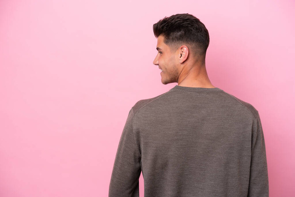 Young caucasian man isolated on pink background in back position and looking side - 写真・画像