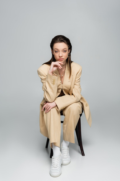 full length of stylish woman in trendy outfit looking at camera while sitting on chair on grey - Photo, image