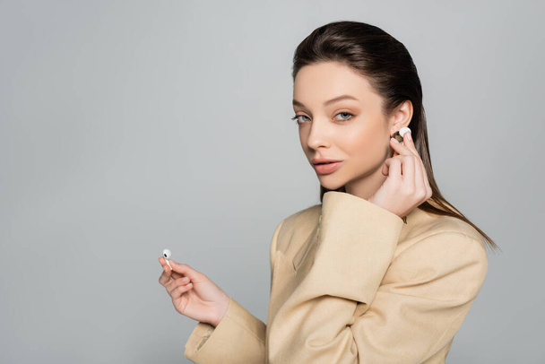young woman in stylish outfit looking at camera while wearing wireless earphones isolated on grey - Photo, Image