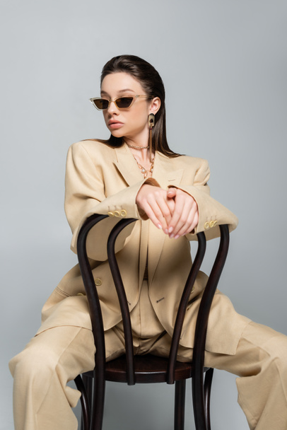 young woman in stylish beige outfit and sunglasses sitting on wooden chair isolated on grey - Photo, Image