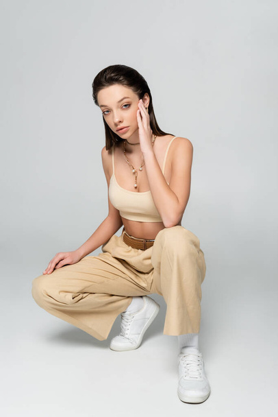 full length of brunette woman in stylish beige outfit and trendy accessories posing while sitting on grey - Photo, image