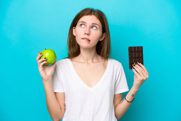 Mladá anglická žena izolovaná na modrém pozadí s pochybnostmi při užívání čokoládové tablety v jedné ruce a jablko v druhé - Fotografie, Obrázek