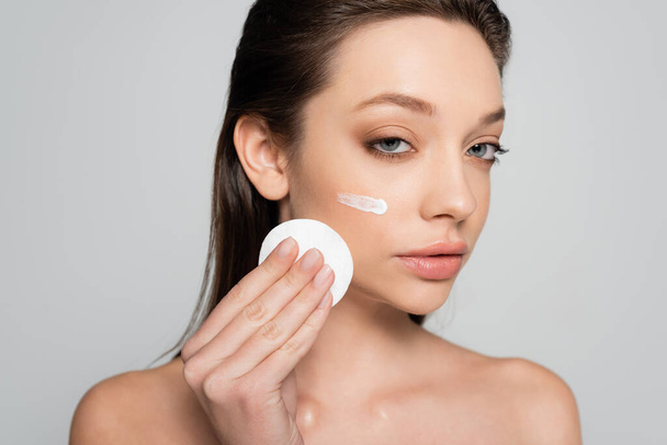 young woman with bare shoulders and cosmetic product on face holding cotton pad isolated on grey - Foto, Imagem