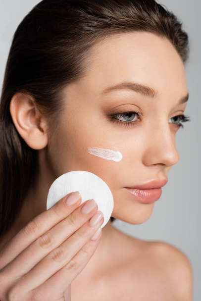 close up of young woman with cosmetic product on face holding cotton pad isolated on grey - Photo, Image